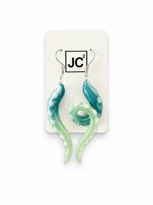Asymmetrical glass tentacle earrings fading from teal to pale green, with white suckers and silver hooks, displayed on a white JC2 card.
