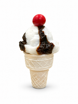 Glass ice cream sculpture with a white scoop, dark brown chocolate topping, and a red cherry in an ivory waffle cone on a white background.
