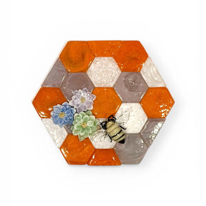 Glass wall sculpture with orange, white, and translucent flower forms arranged in a hexagonal composition on a white background.
S