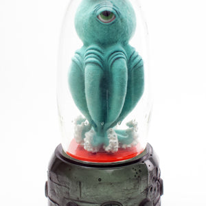 Nomad mixed-media sculpture featuring a teal, one-eyed tentacled figure inside a clear glass enclosure with orange accents and a gray base.
