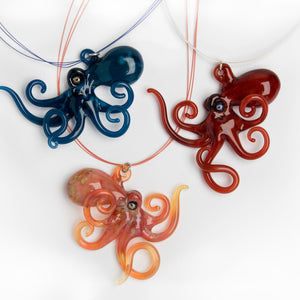 
Three flameworked glass octopus necklaces in blue, red, and coral, with curling tentacles, displayed on a white background.
Sent f
