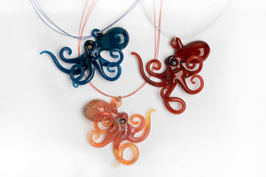 
Three flameworked glass octopus necklaces in blue, red, and coral, with curling tentacles, displayed on a white background.
Sent f