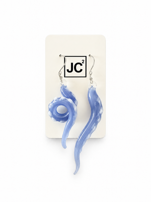 Asymmetrical periwinkle-blue glass tentacle earrings with white suckers, one curled and one long, on silver hooks displayed on a white JC2 card.
