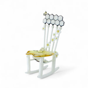 White glass rocking chair sculpture with a honeycomb backrest, a small bee, white flowers with yellow centers, and a translucent golden-yellow seat.
