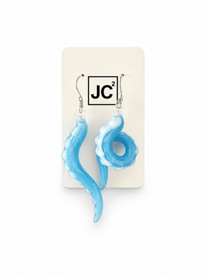 Asymmetrical sky-blue glass tentacle earrings with white suckers, one long and one curled, on silver hooks displayed on a white JC2 card.
