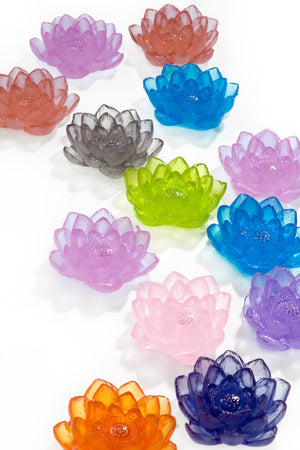 Cast glass water lilies with layered translucent petals in pink, purple, blue, coral, orange, gray, and lime green, arranged on a white background.

