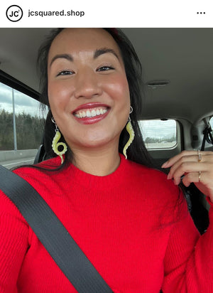 Smiling woman in a red sweater wearing asymmetrical yellow-green glass tentacle earrings, showing their length when worn.

